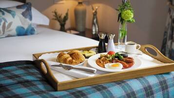A breakfast tray with croissants, eggs, beans, and a fruit salad on a bed with a plaid blanket.