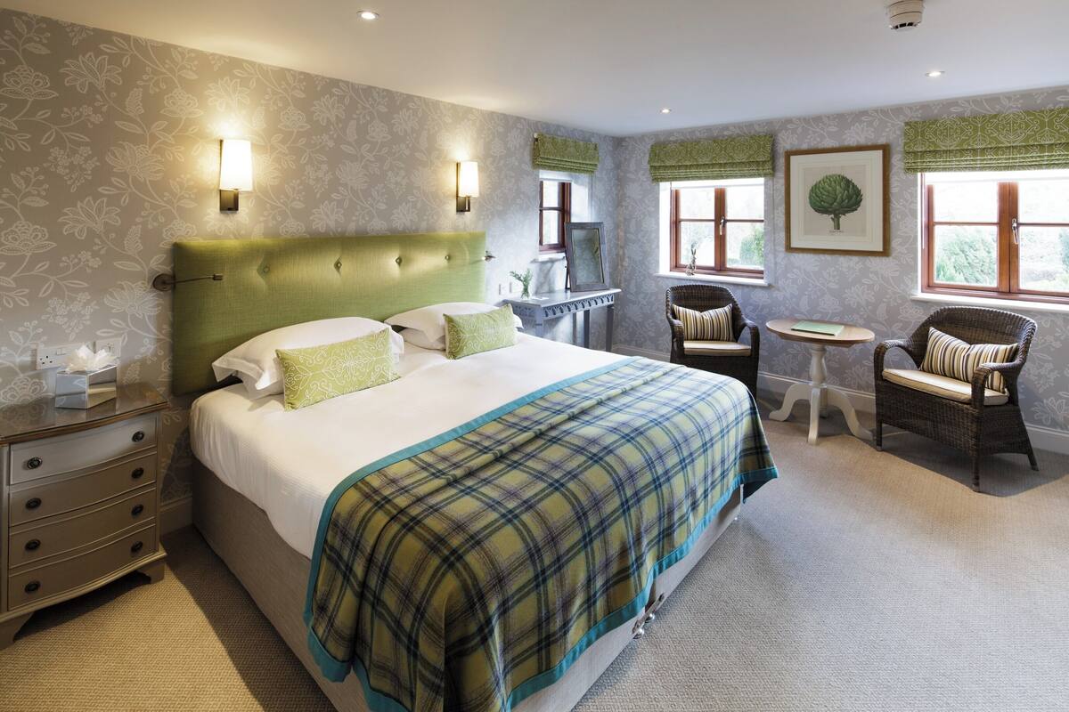 Une chambre d’hôtel avec un grand lit, une table de chevet, une chaise, une petite table et une fenêtre avec des rideaux.