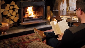 Una persona leyendo un libro en una habitación acogedora con chimenea, una mesa con una taza y un plato, y una pila de leña.