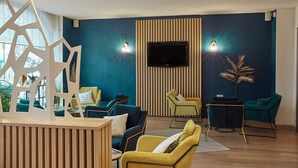 Un área de descanso moderna con un mostrador de recepción de madera, asientos en tonos verde azulado y amarillo, un televisor de pantalla plana montado en una pared a rayas y dos luces empotradas.