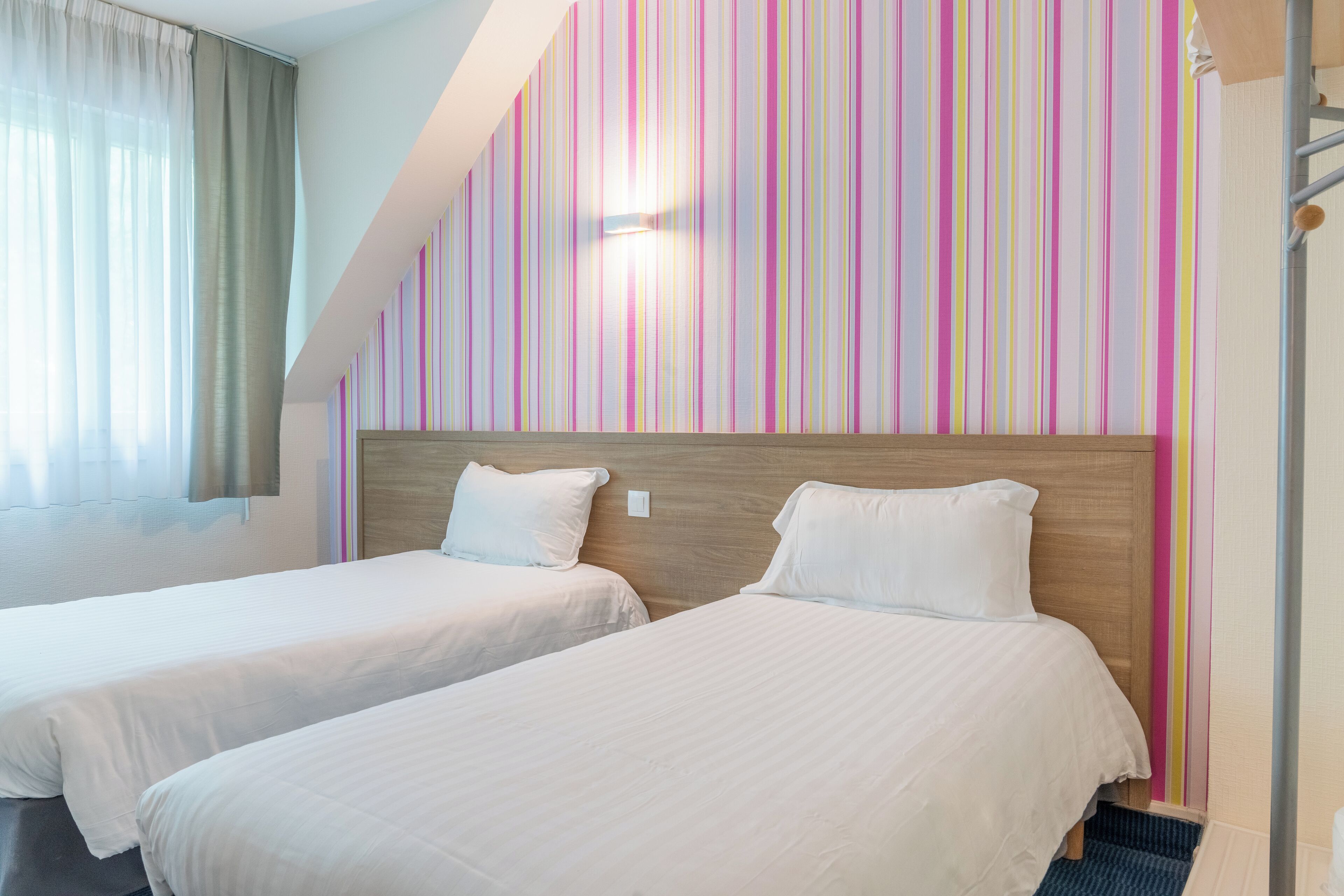 Habitación de hotel con dos camas, cabecero de madera, pared a rayas y una ventana con cortinas.
