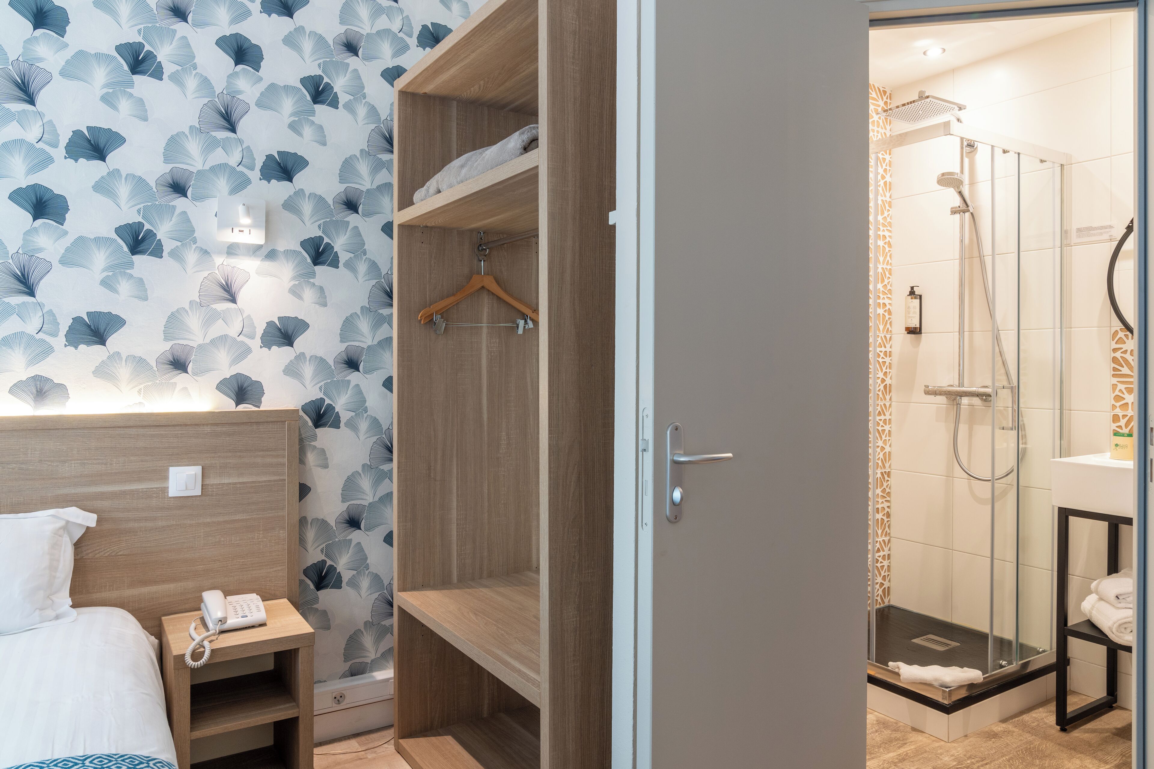 A hotel room with a wooden headboard, a bedside phone, a wardrobe with a hanging rack, and a shower area with a glass door.