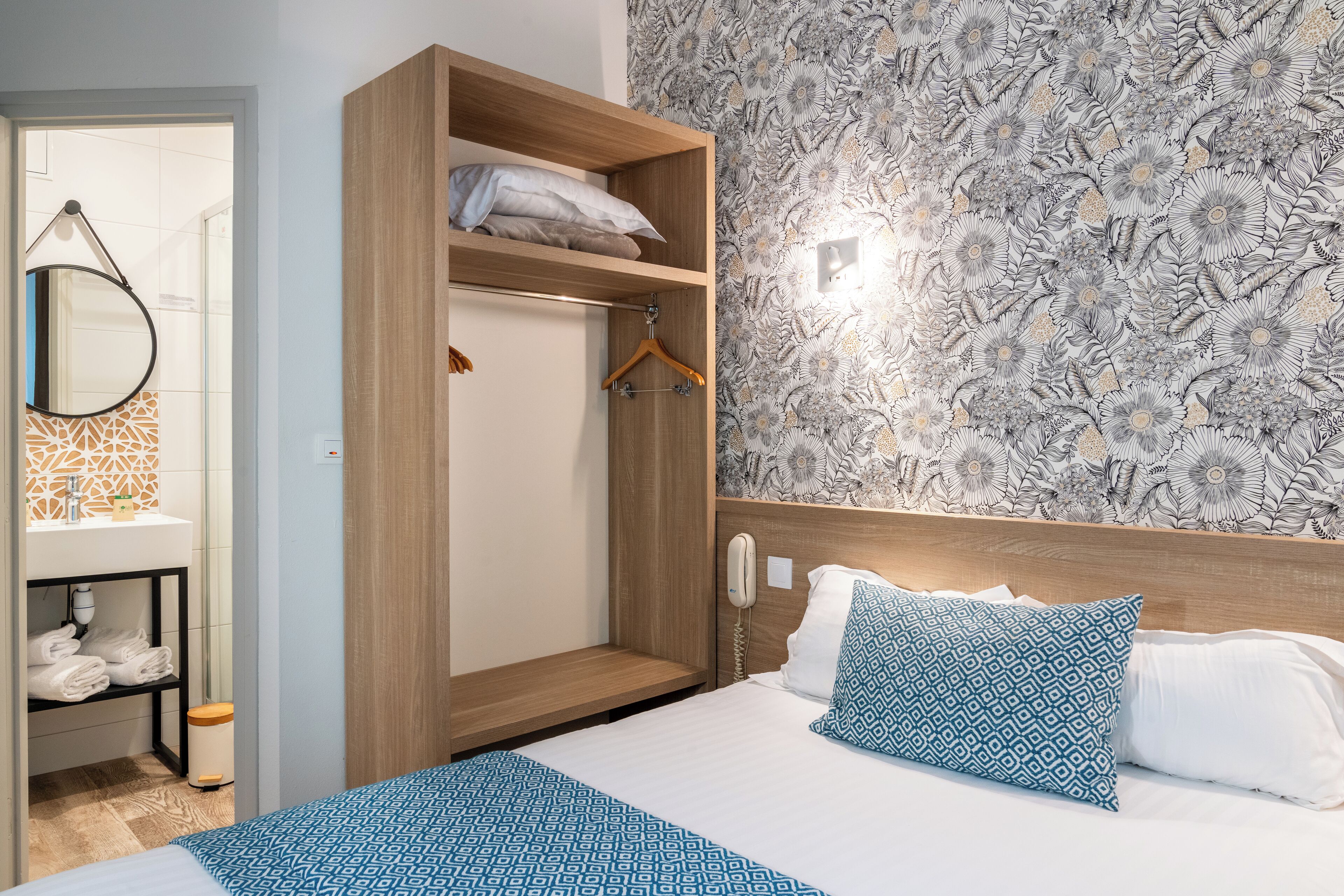 Quarto de hotel com guarda-roupa de madeira, cama com roupa de cama estampada, casa de banho com espelho e suporte de toalhas, e papel de parede floral.