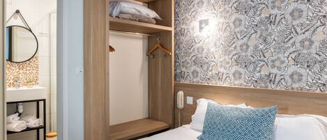 Habitación de hotel con armario de madera, cama con ropa de cama estampada, baño con espejo y toallero, y papel tapiz floral.