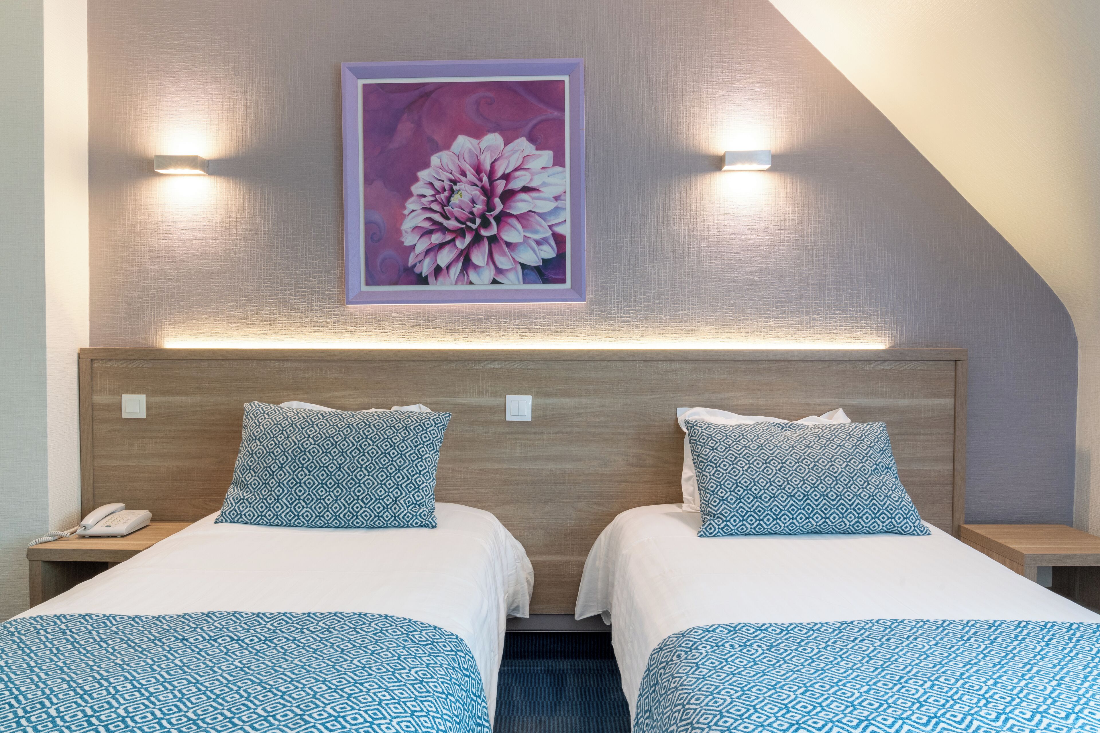 A hotel room with two beds, a wooden headboard, a wall-mounted painting, and a phone on the nightstand.