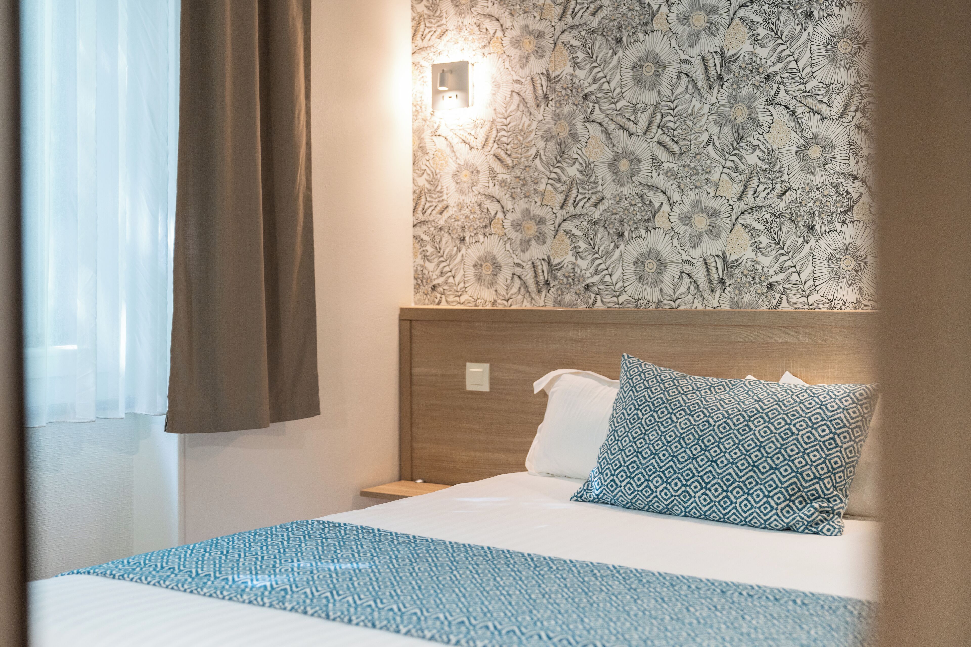A hotel room with a bed, a nightstand, a wall-mounted light, and a patterned wallpaper.