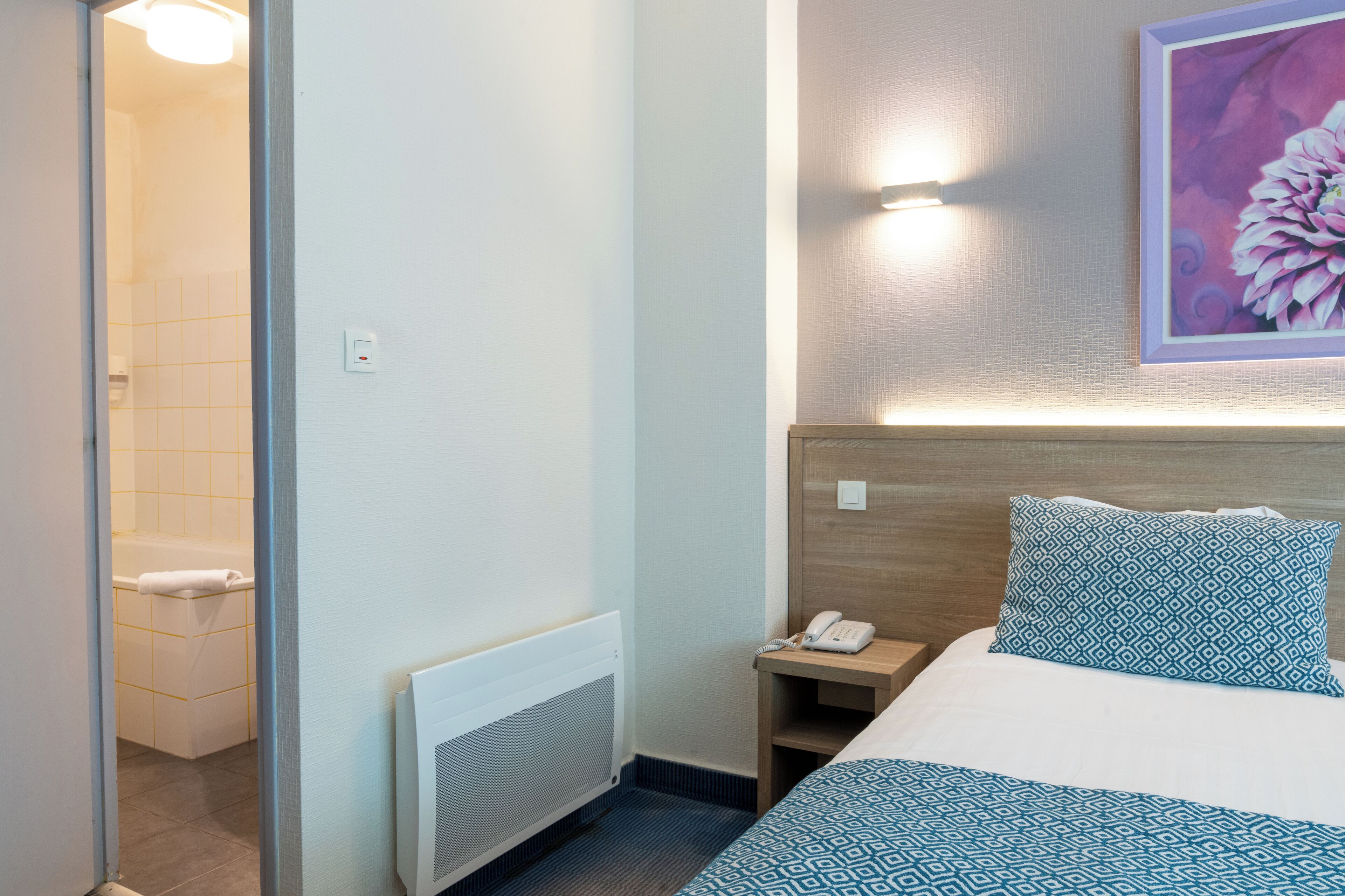 A hotel room with a bed, a nightstand with a phone, a radiator, and a painting on the wall.