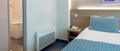 A hotel room with a bed, a nightstand with a phone, a radiator, and a painting on the wall.