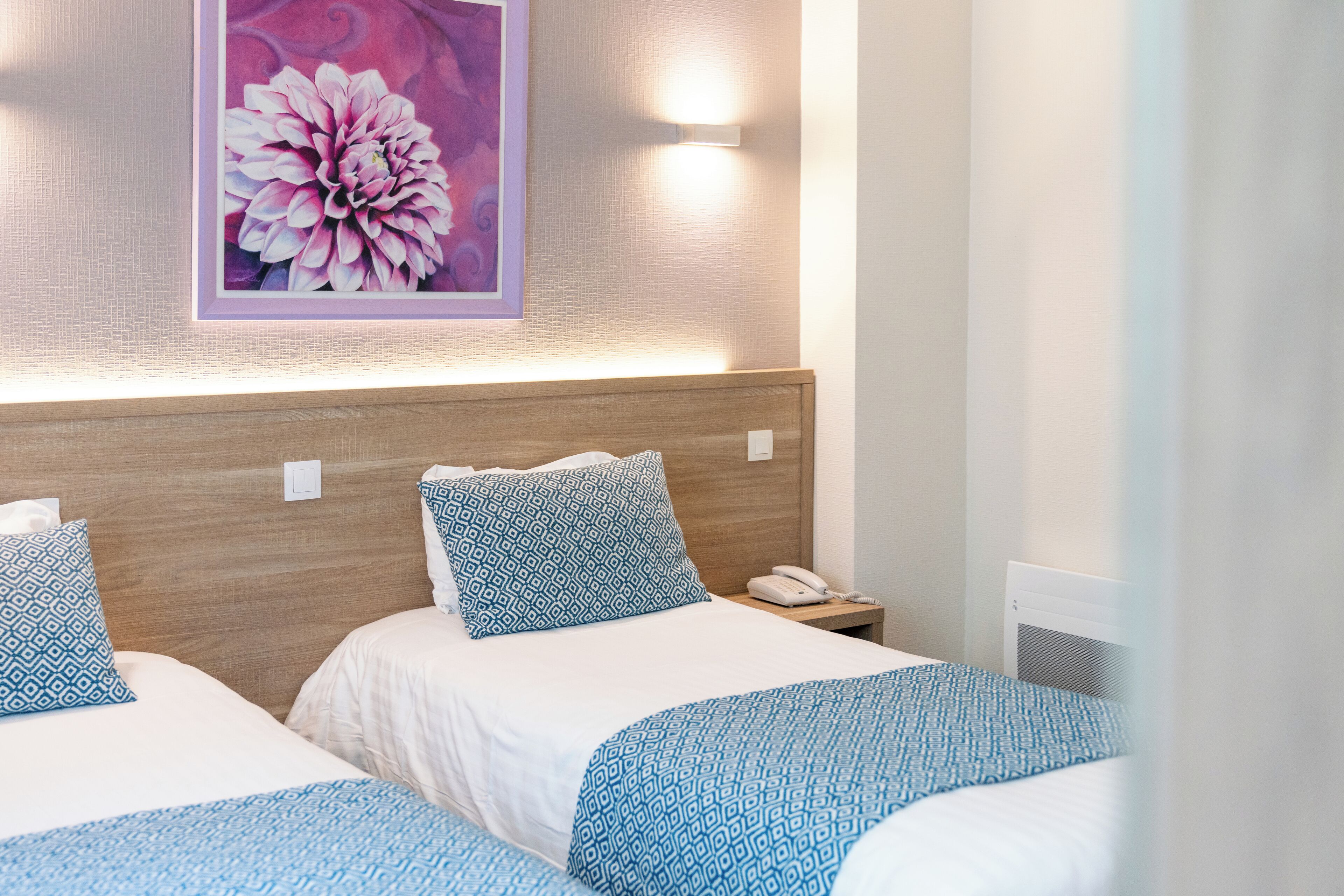 A hotel room with two beds, each with a patterned bedspread, a wooden headboard, a bedside table with a phone, and a wall-mounted light.