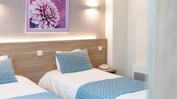 A hotel room with two beds, each with a patterned bedspread, a wooden headboard, a bedside table with a phone, and a wall-mounted light.