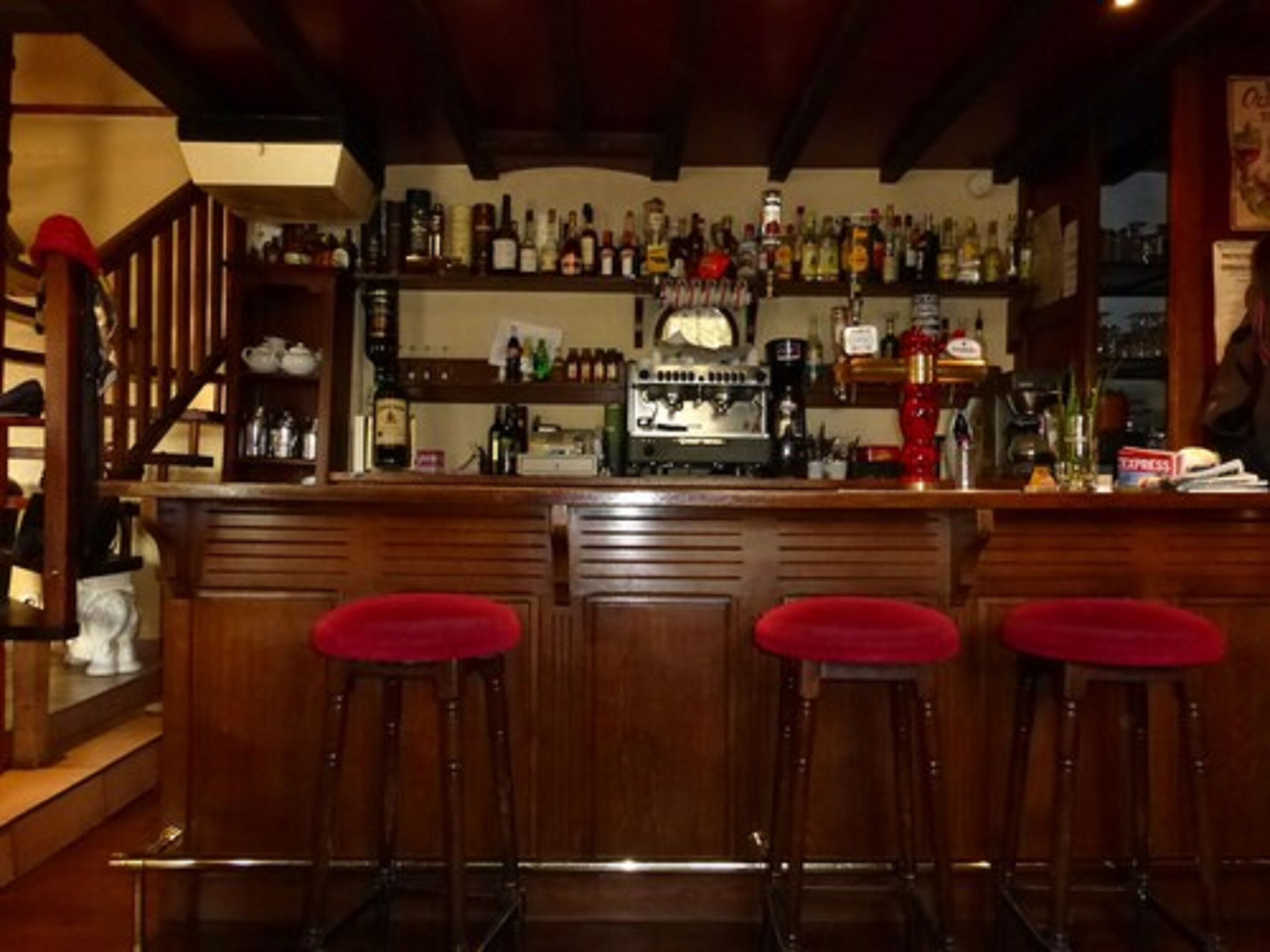 A bar area with wooden stools, a bar counter, and shelves stocked with various bottles.