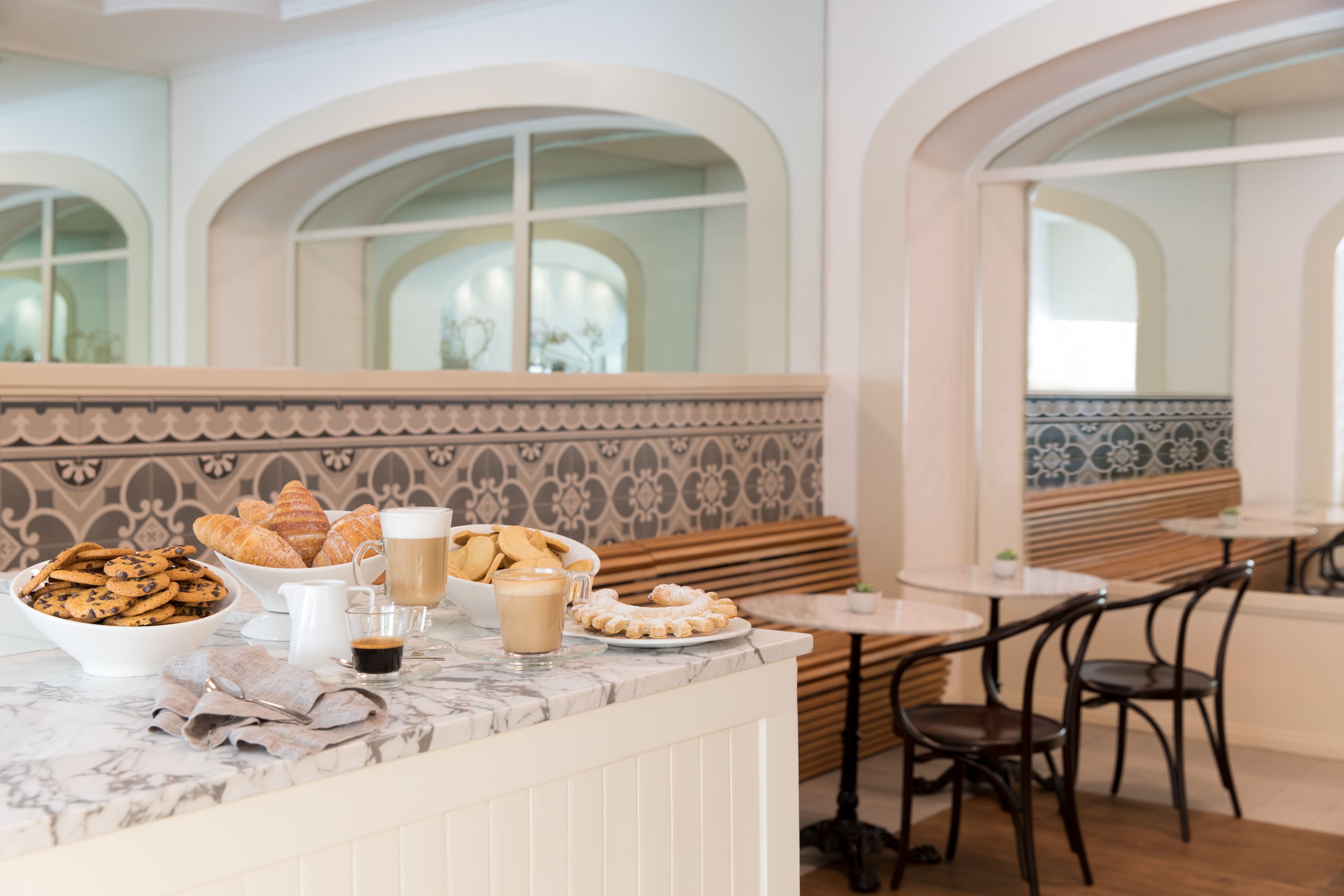 Un desayuno con una encimera de mármol, un bol de galletas, pasteles y café. Se observan bancos y mesas de madera al fondo.