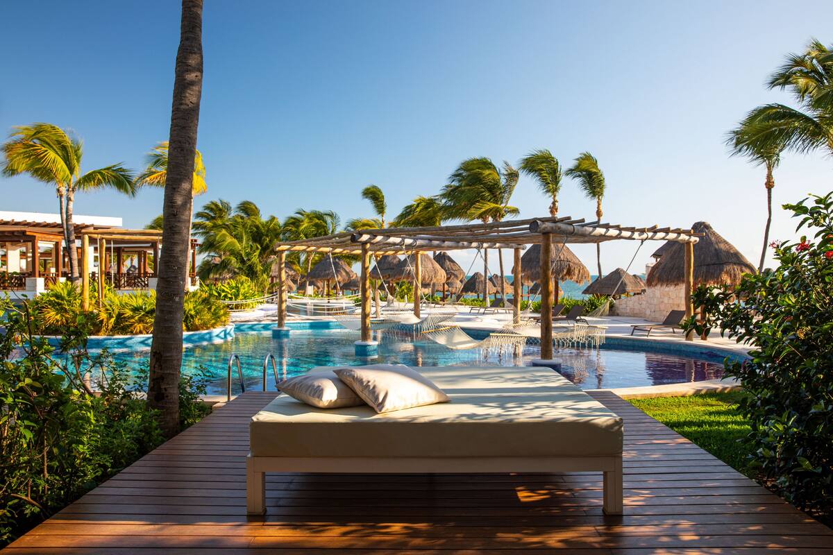 A poolside area with a wooden deck, lounge chairs, and a thatched-roof structure.