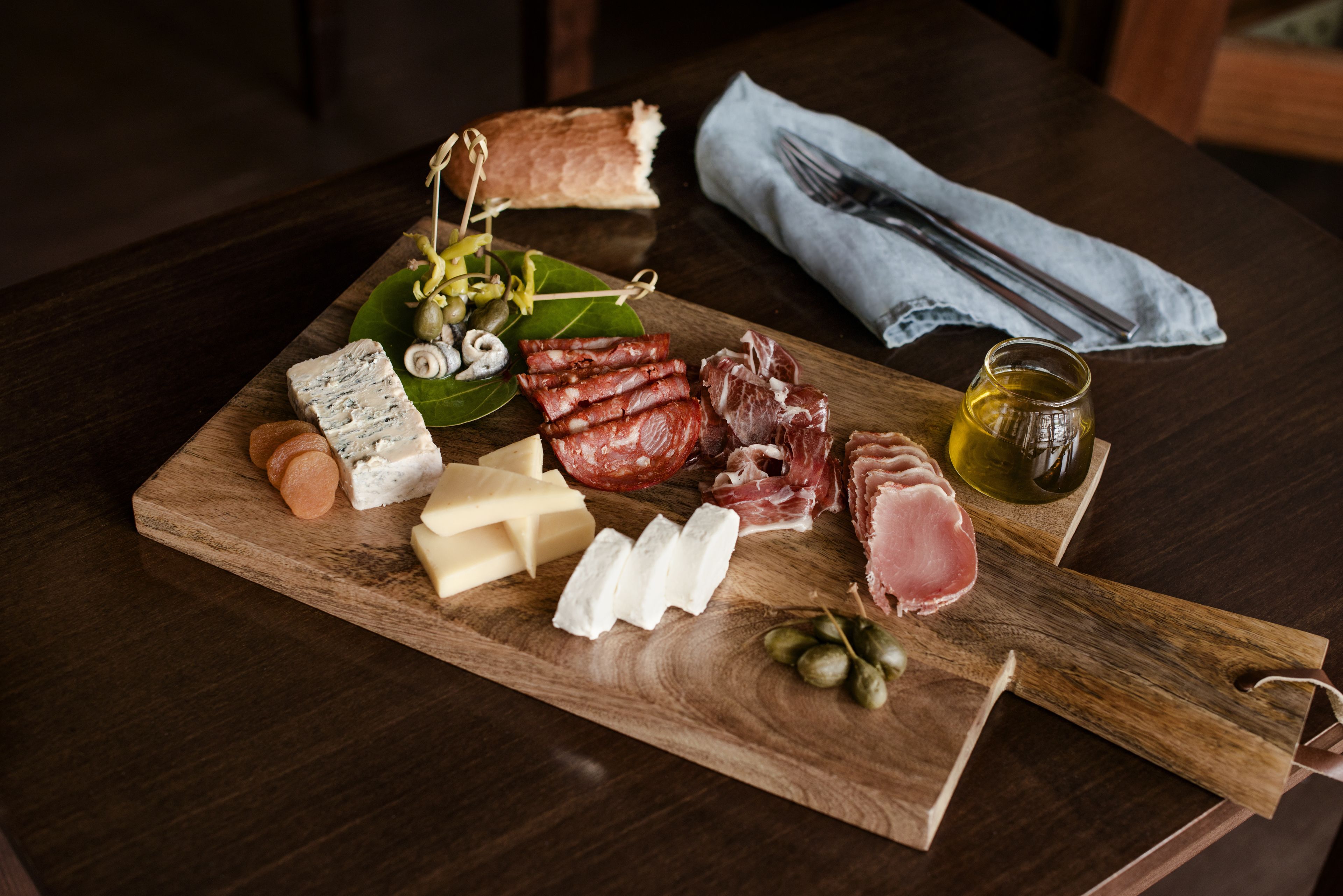 A wooden board with various cheeses, cured meats, olives, and a bottle of olive oil.