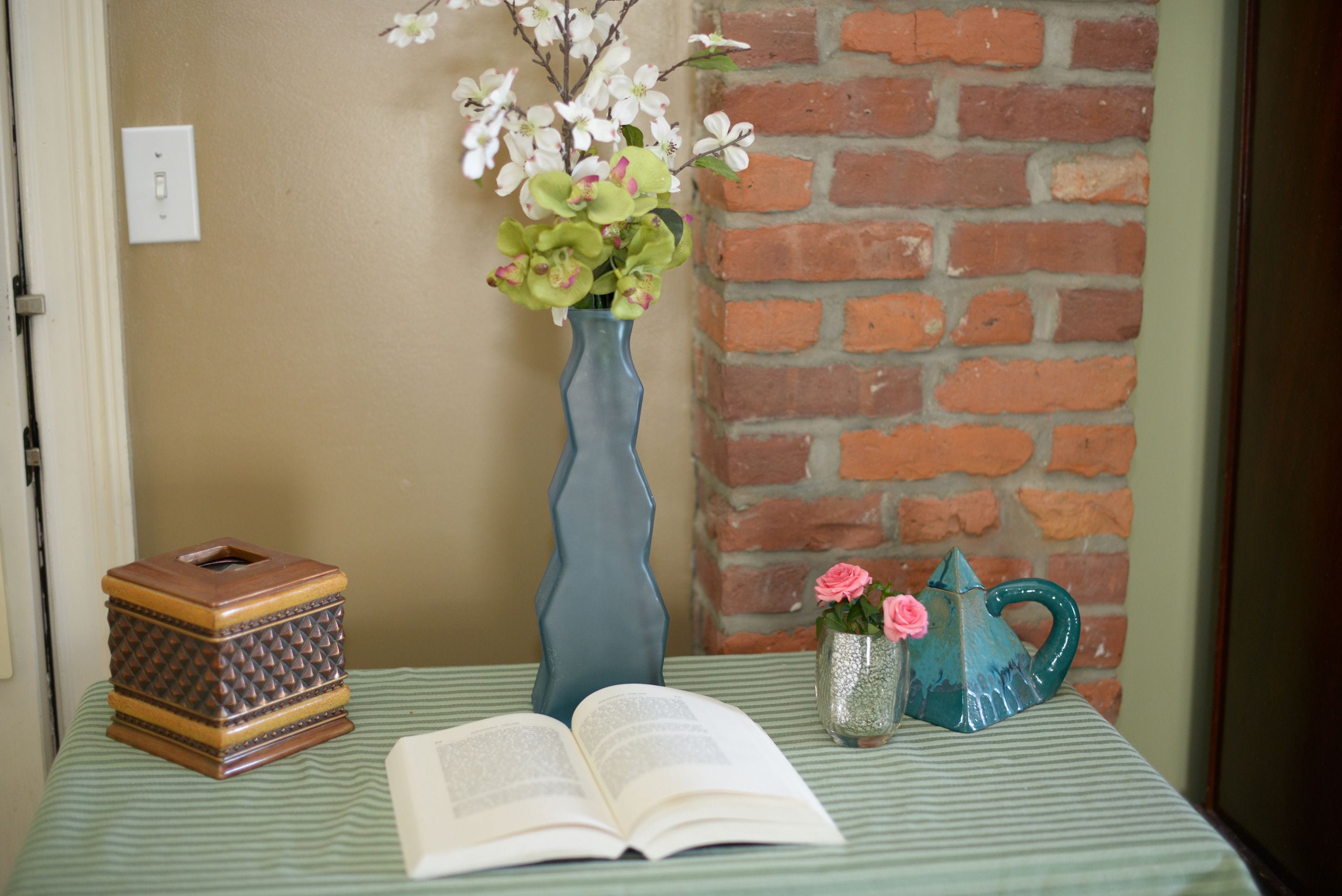 Une pièce avec un mur en briques, une table sur laquelle se trouve un livre, un vase rempli de fleurs, une boîte décorative et une théière.