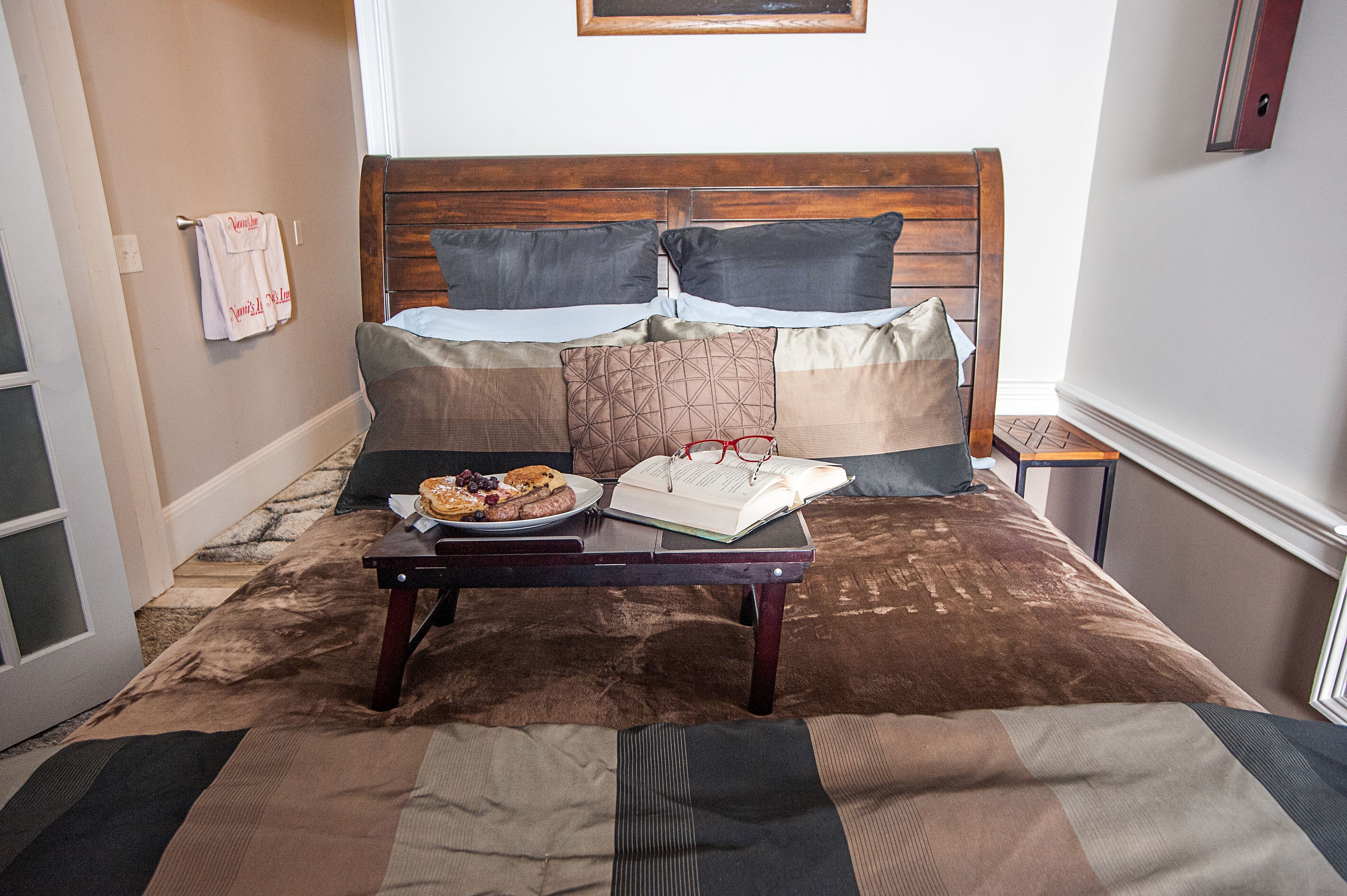 A neatly made bed with a wooden headboard, a bedside table with a book and a plate of food, and a towel hanging on a hook.