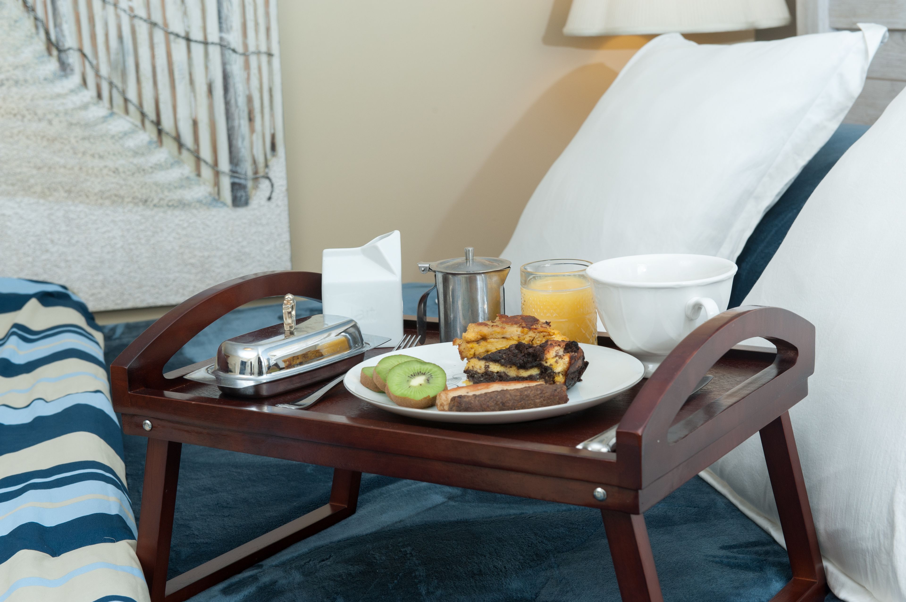 A breakfast tray with a slice of cake, kiwi, orange juice, and a coffee pot on a bed with a pillow and a blanket.