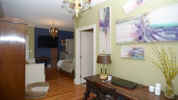 A room with a wooden floor, a desk with a lamp, a laptop, and a vase with flowers. There is a bed with a headboard in the background.