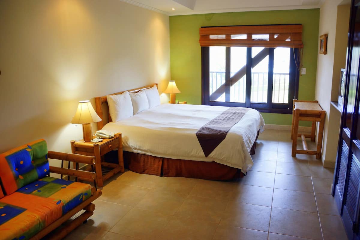 A hotel room with a bed, a wooden nightstand, a colorful sofa, a window with a view, and a small wooden table.