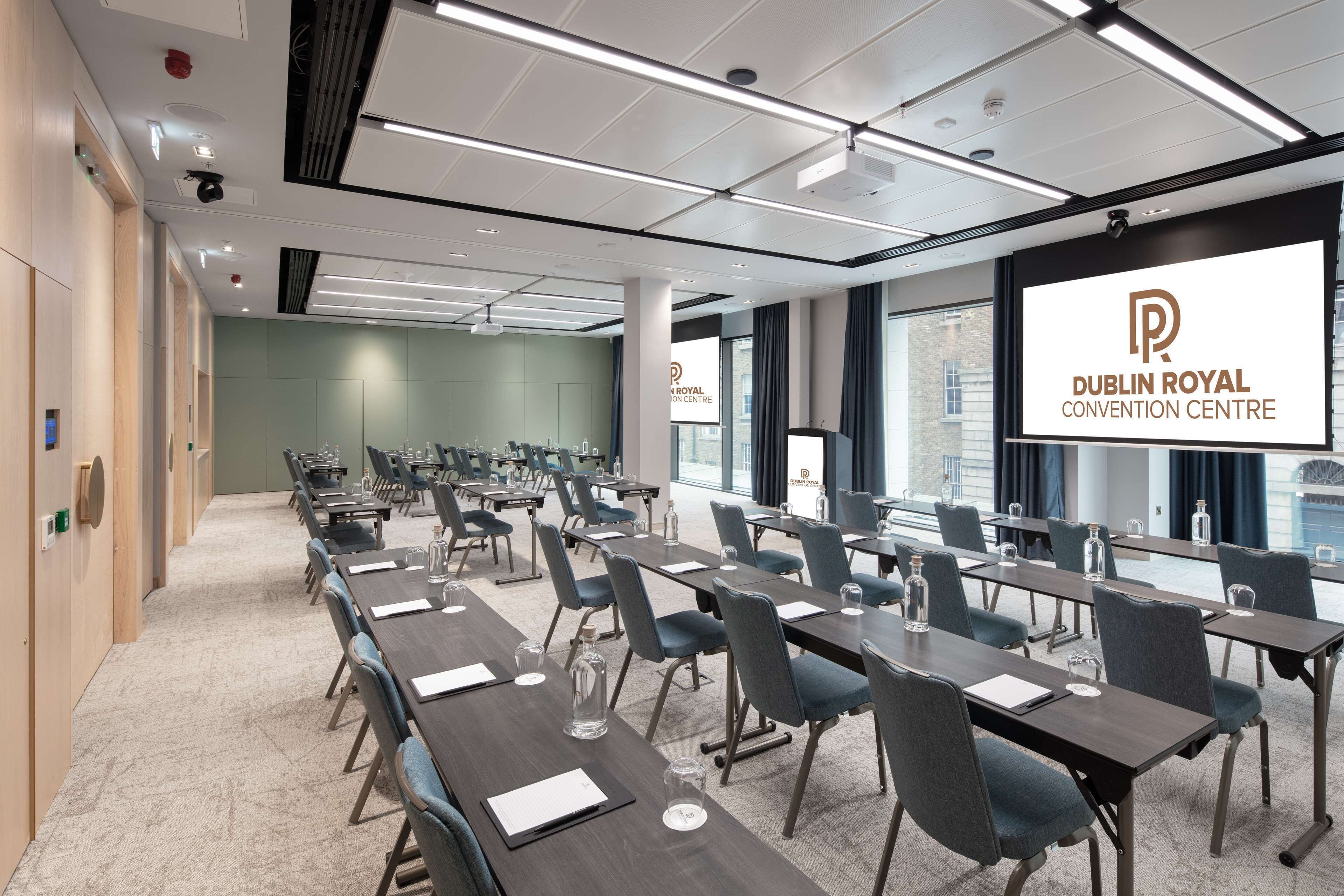 Een congrescentrum met lange tafels, stoelen en een groot scherm waarop het logo van het Dublin Royal Convention Centre staat.