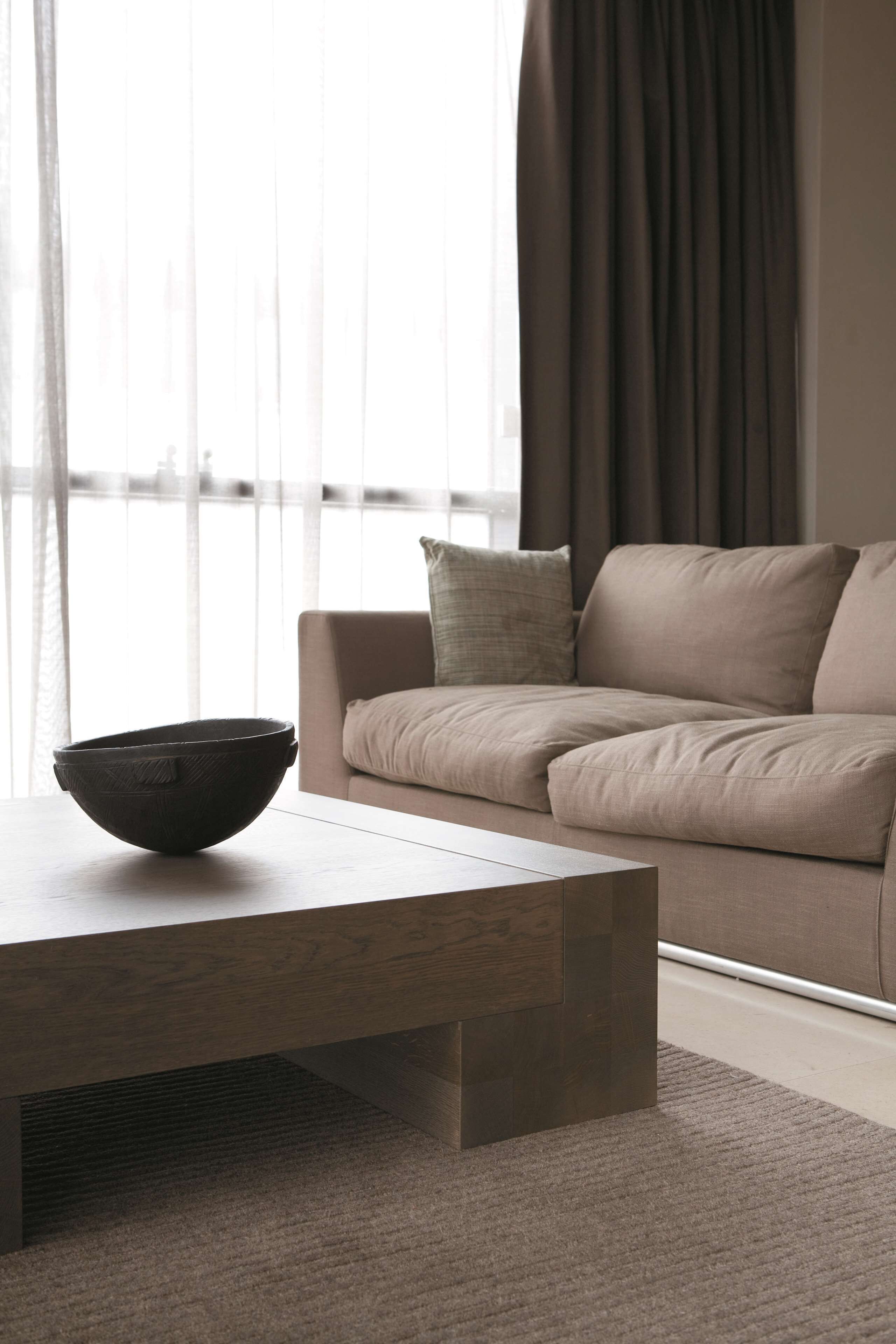 A modern living room with a beige sofa, a wooden coffee table, and a large window with sheer curtains.