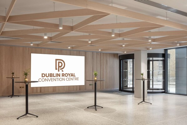 A spacious convention center with a modern design, featuring a large screen displaying the Dublin Royal Convention Centre logo, and three small tables with potted plants.