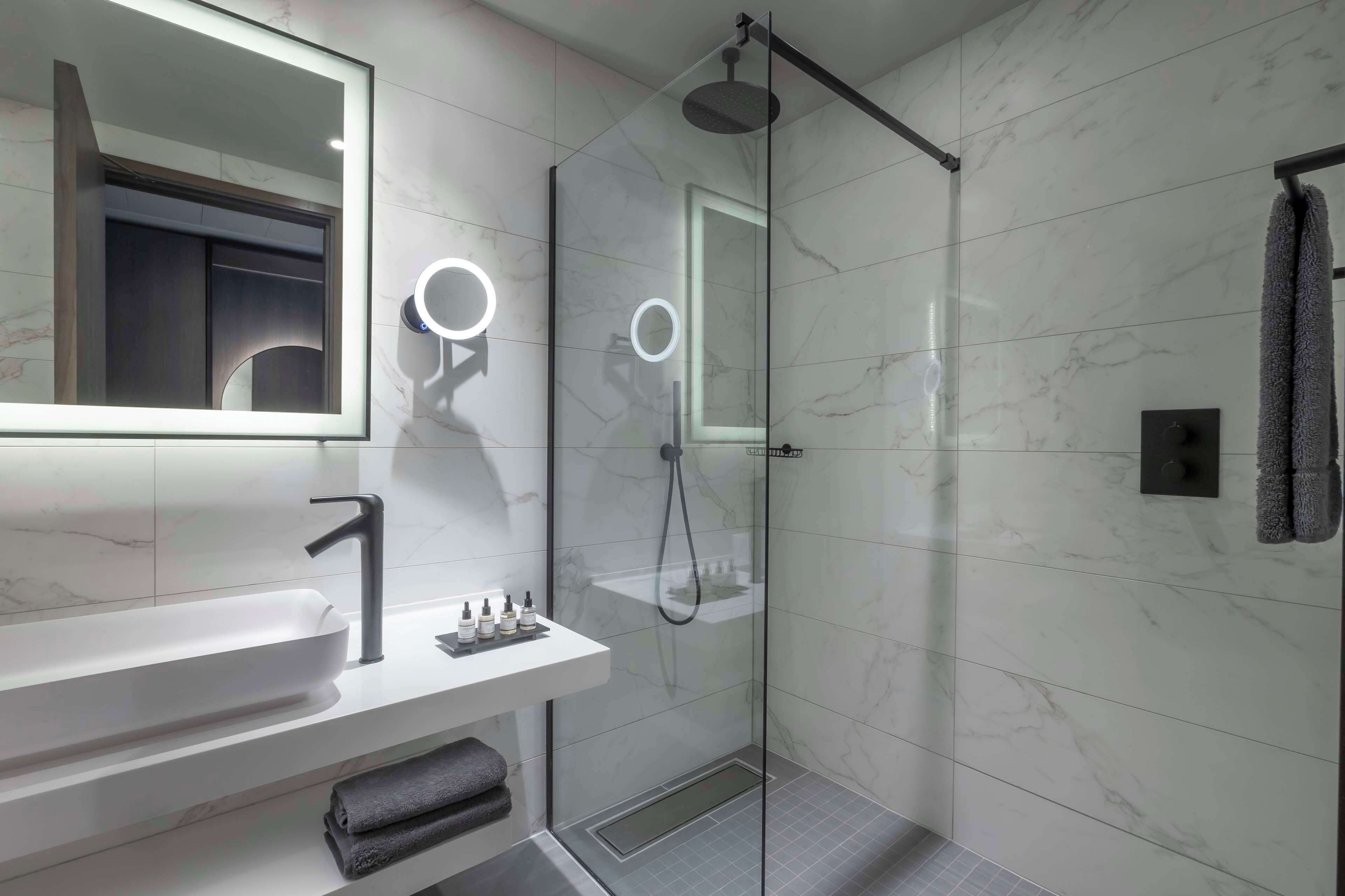Un baño moderno con ducha de vidrio, un tocador blanco con grifo elegante y un espejo grande con iluminación integrada.