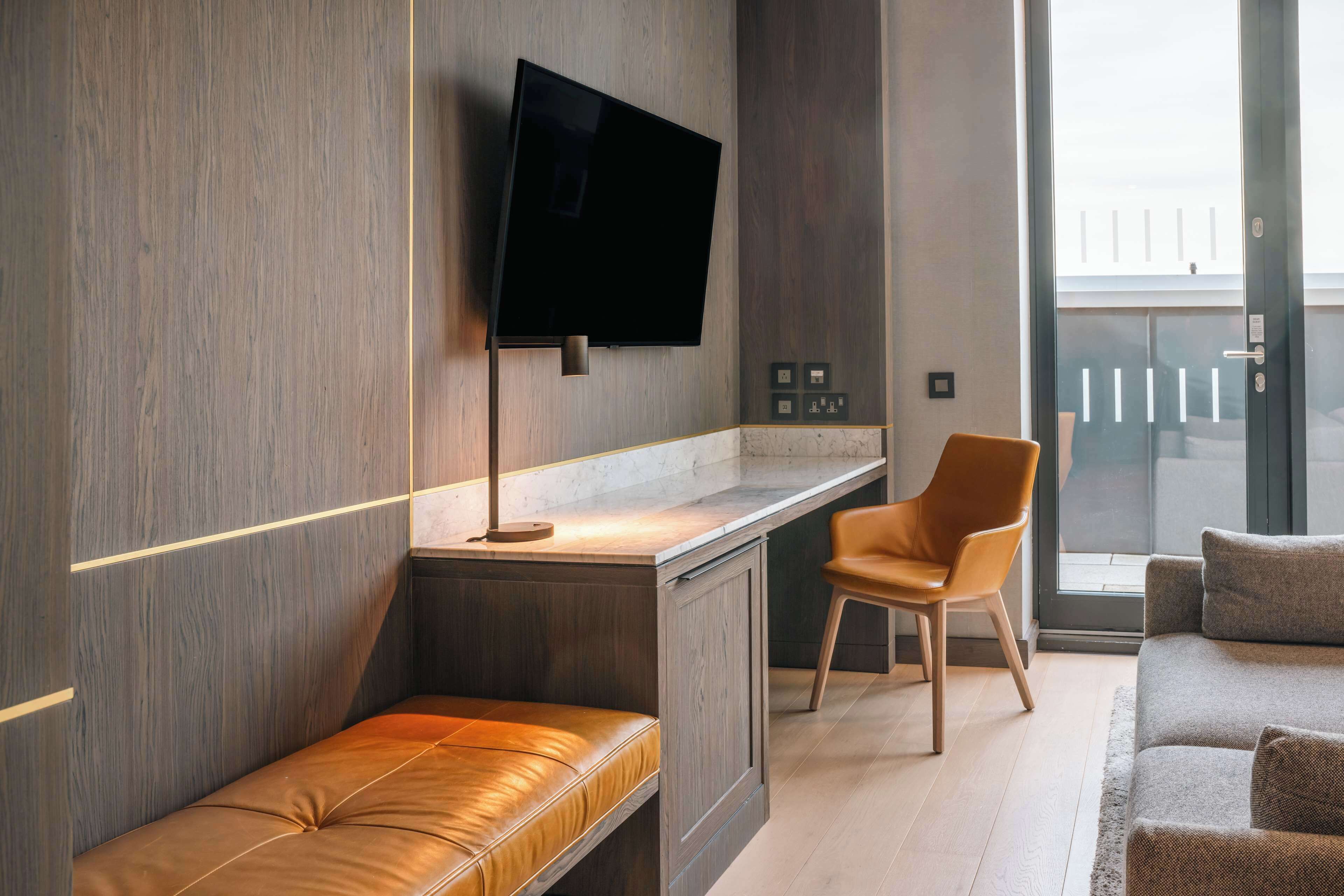 A modern room with a wooden wall, a flat-screen TV mounted above a desk, a leather bench, a brown chair, and a sofa.