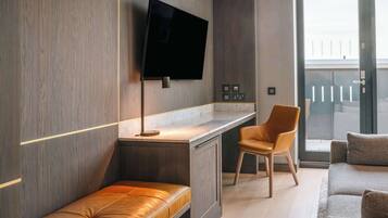 A modern room with a wooden wall, a flat-screen TV mounted above a desk, a leather bench, a brown chair, and a sofa.