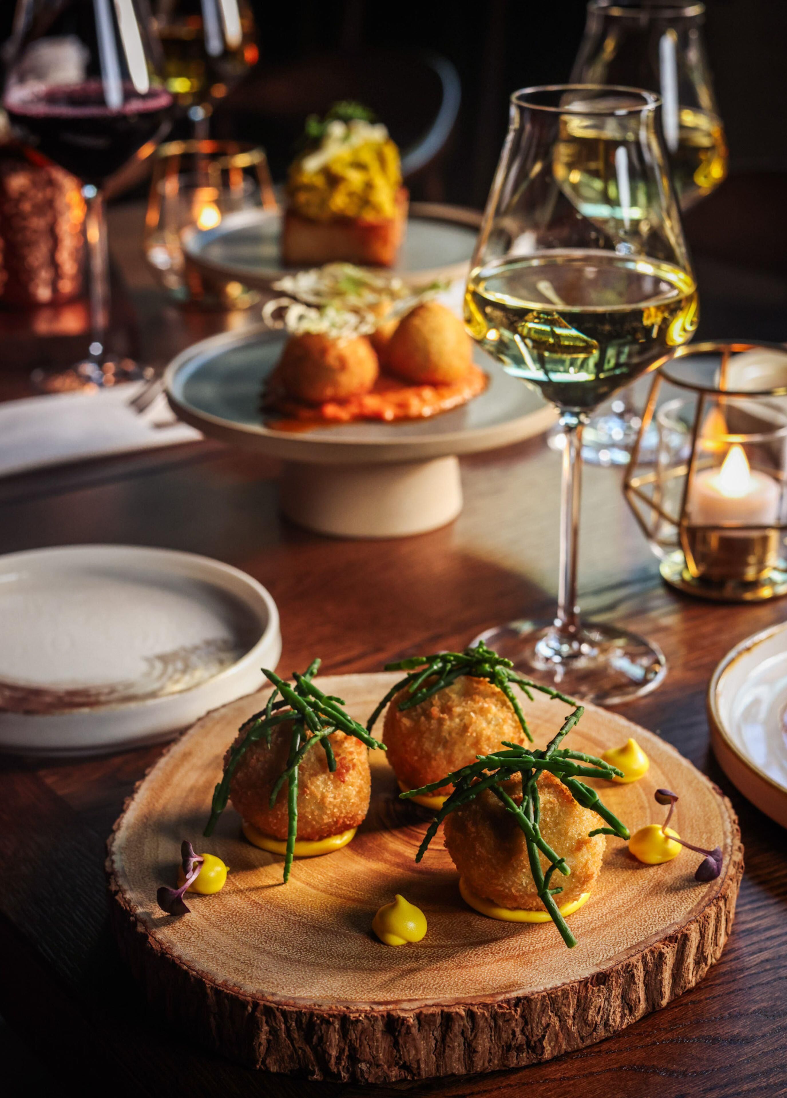 Una mesa de madera con un plato de comida, una copa de vino blanco y una vela.