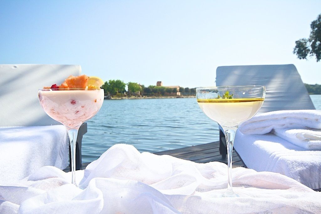 Two drinks on a table by a lake with towels and a view of a building in the background.