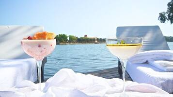 Two drinks on a table by a lake with towels and a view of a building in the background.