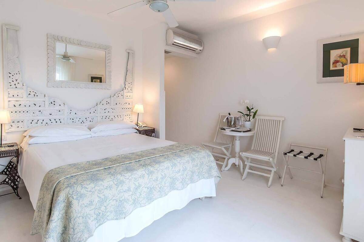 A bedroom with a bed, a chair, a small table, a lamp, an air conditioner, and a mirror.