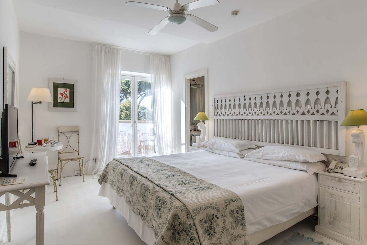 A bedroom with a large bed, a desk, a chair, a ceiling fan, and a window with curtains.