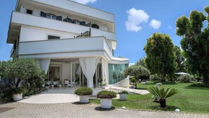 A modern multi-story building with a curved entrance, a covered outdoor seating area, and a well-maintained garden with potted plants and a lawn. - Il San Francesco Charming Hotel (Sabaudia)