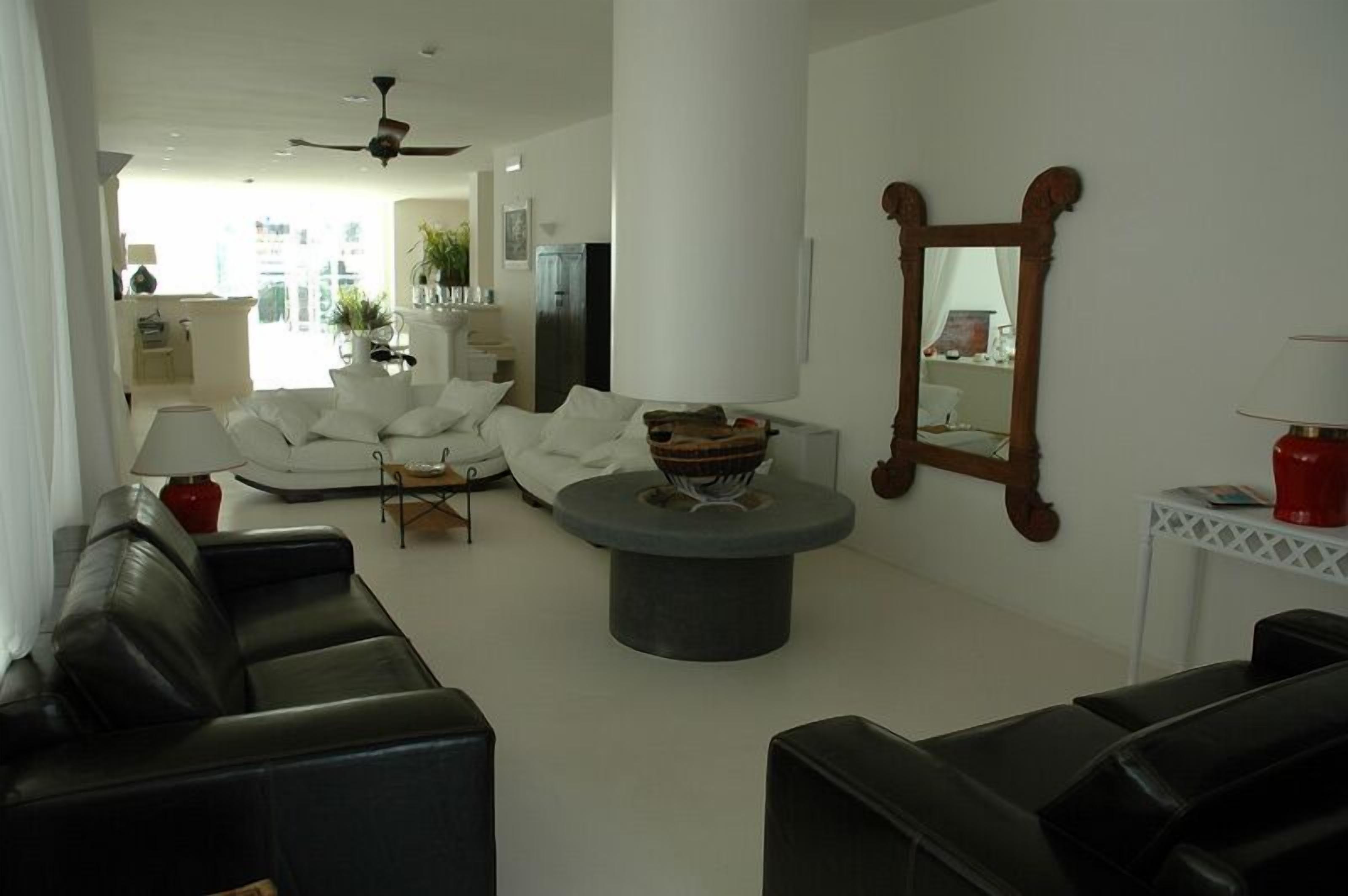 A modern living room with a black leather sofa, a round coffee table, and a white couch. There is a wooden mirror on the wall and a red lamp on a side table.