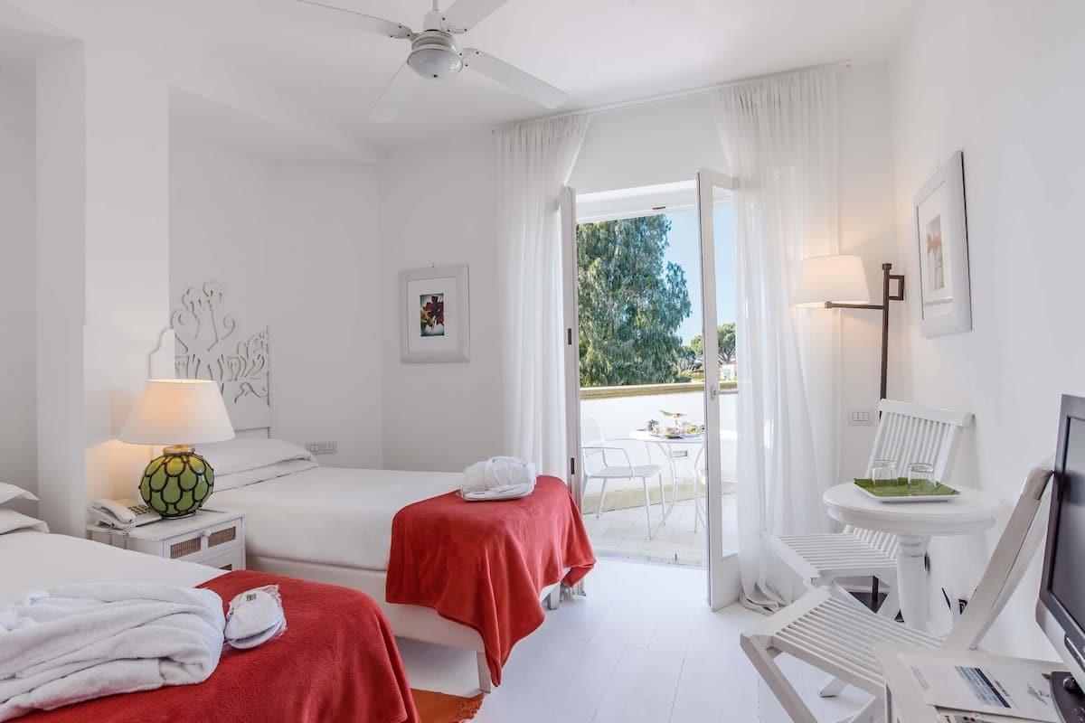A hotel room with two beds, a desk with a computer, a chair, a table with a lamp, and a view of the outdoors.