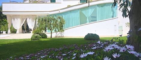 Ein modernes, zweistöckiges Gebäude mit weißer Fassade, einem großen Glasfenster und einem gepflegten Garten mit violetten Blumen.