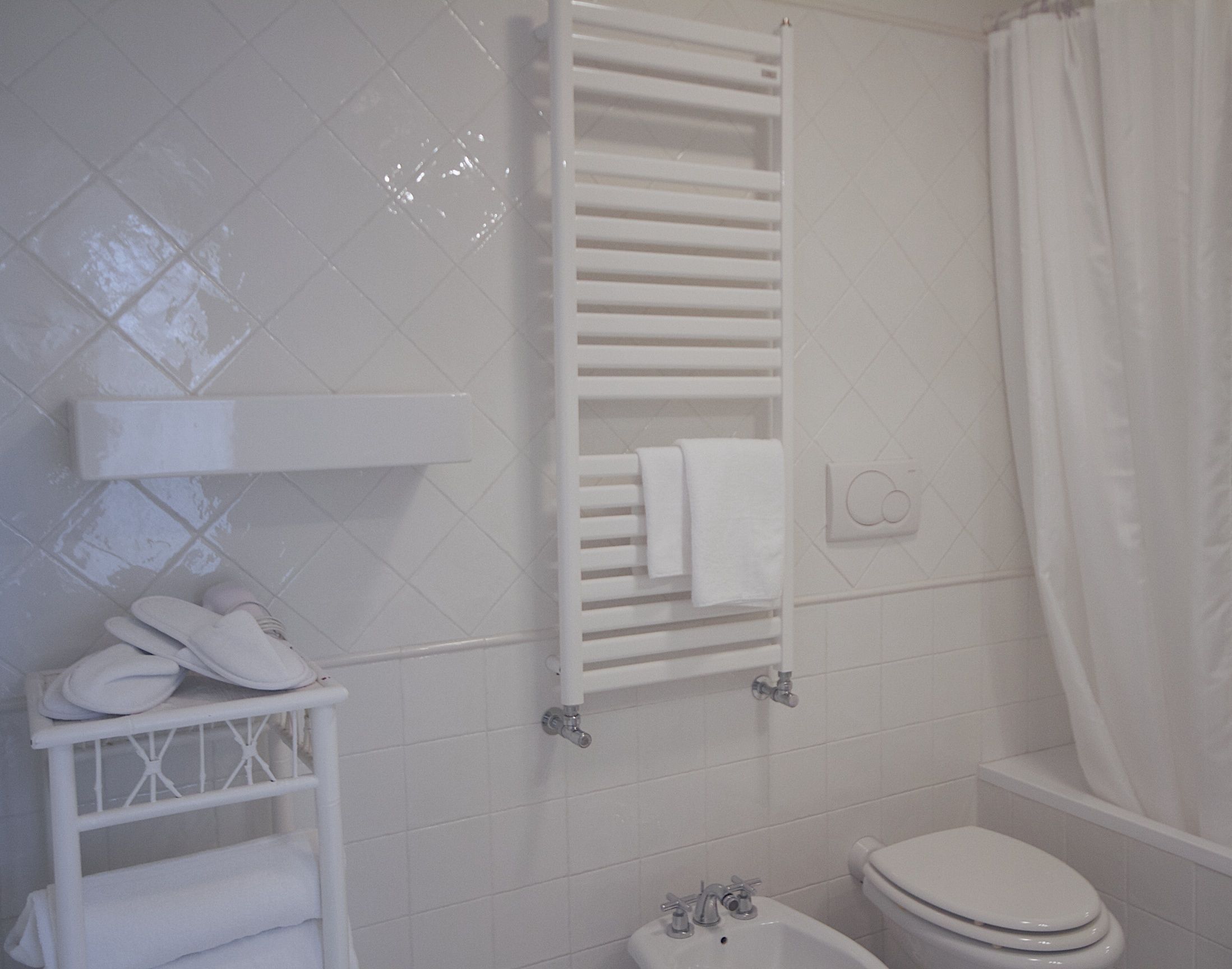 Un baño con una bañera blanca, un inodoro blanco y un lavamanos blanco. Hay un perchero con toallas y un par de zapatillas en un estante.