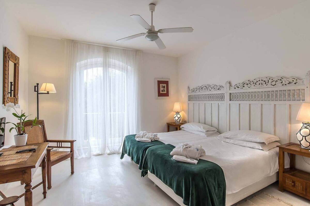 A hotel room with a bed, two bedside tables, a desk with a chair, a ceiling fan, and a window with curtains.