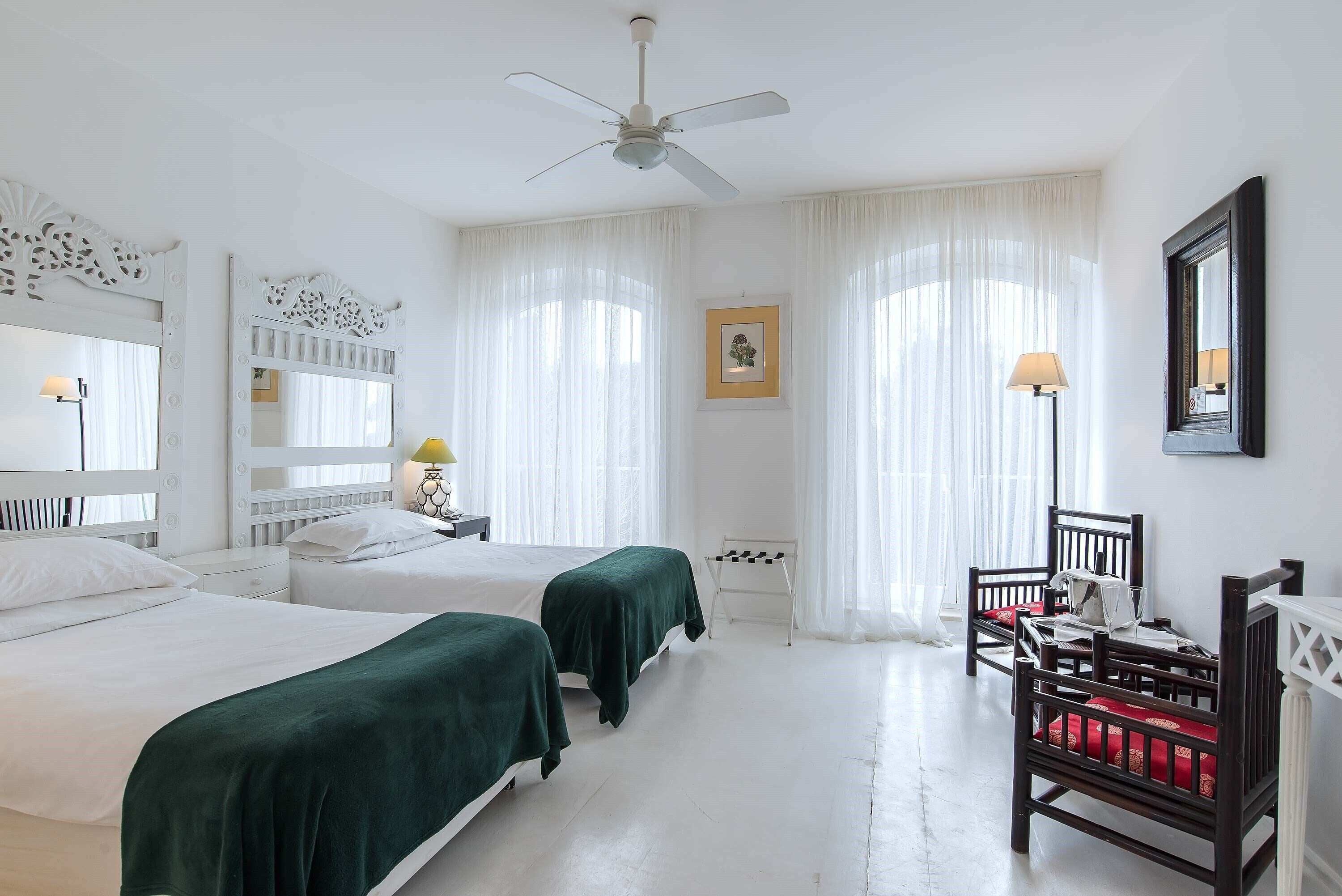 Habitación de hotel con dos camas, ventilador de techo, escritorio con silla y un gran ventanal con cortinas.