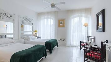 Habitación de hotel con dos camas, ventilador de techo, escritorio con silla y un gran ventanal con cortinas.