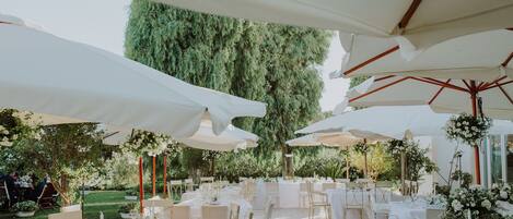 An outdoor dining area with white tables and chairs, covered by large white umbrellas, surrounded by greenery.