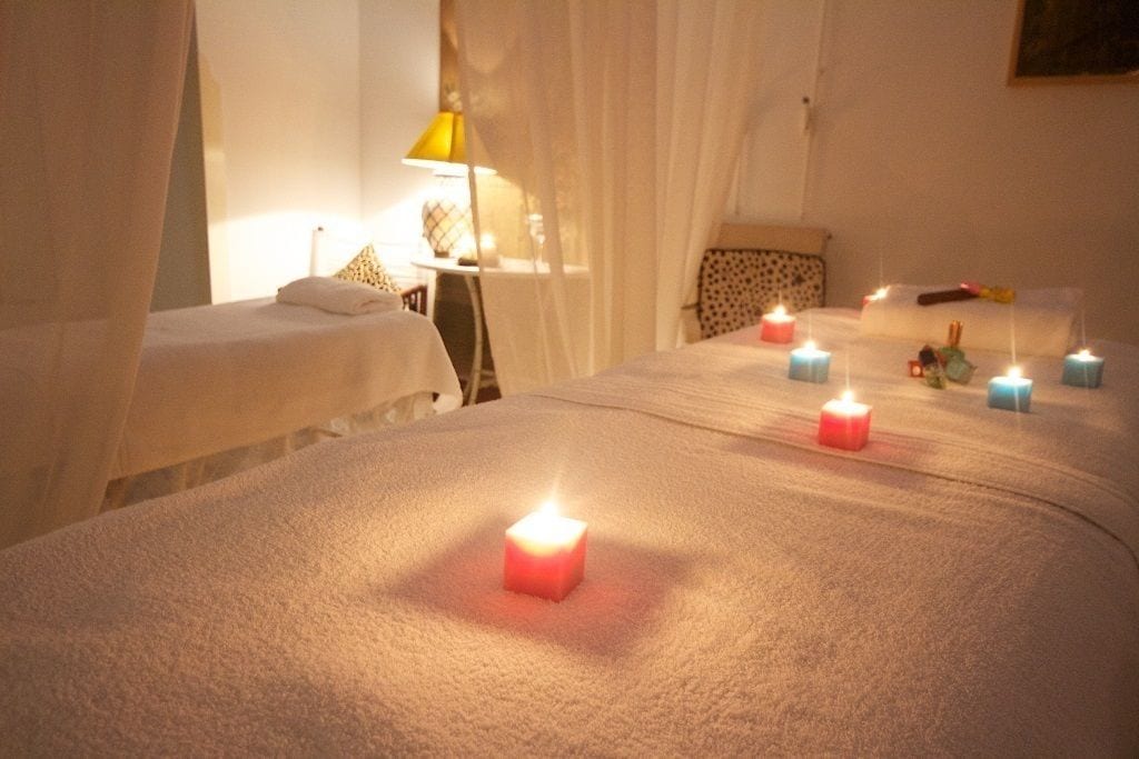 A room with two beds, each with a white bedspread and a small bedside table with a lamp. There are candles on the beds and a painting on the wall.