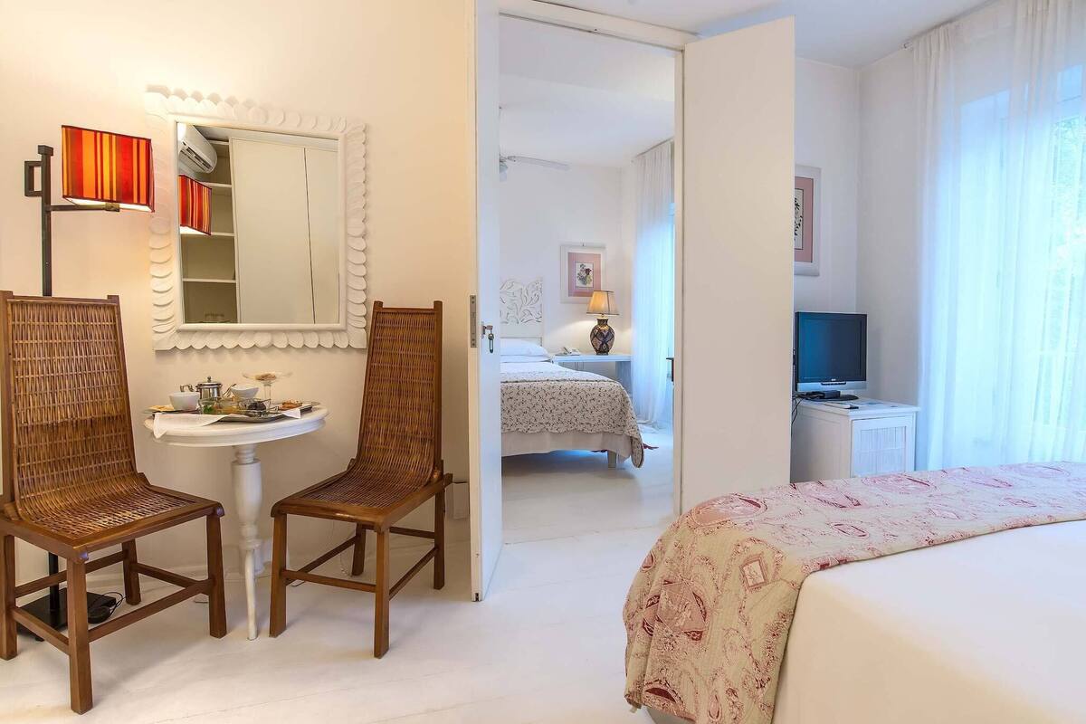 A hotel room with a bed, two wicker chairs, a small round table with a lamp, a mirror, a wardrobe, a television, and a bathroom visible through an open door.