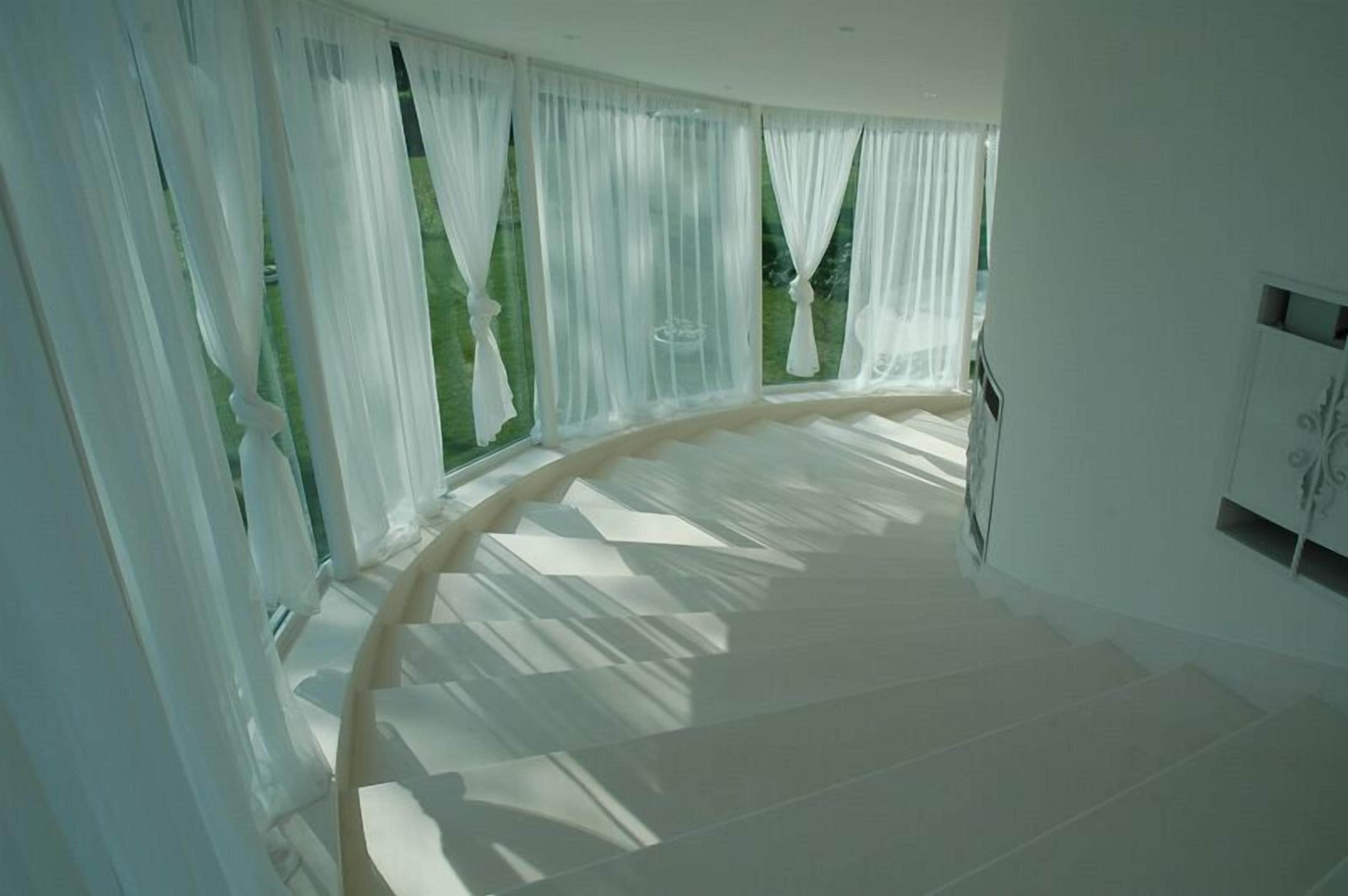 A room with a curved ceiling, large windows with sheer curtains, and a white tiled floor.