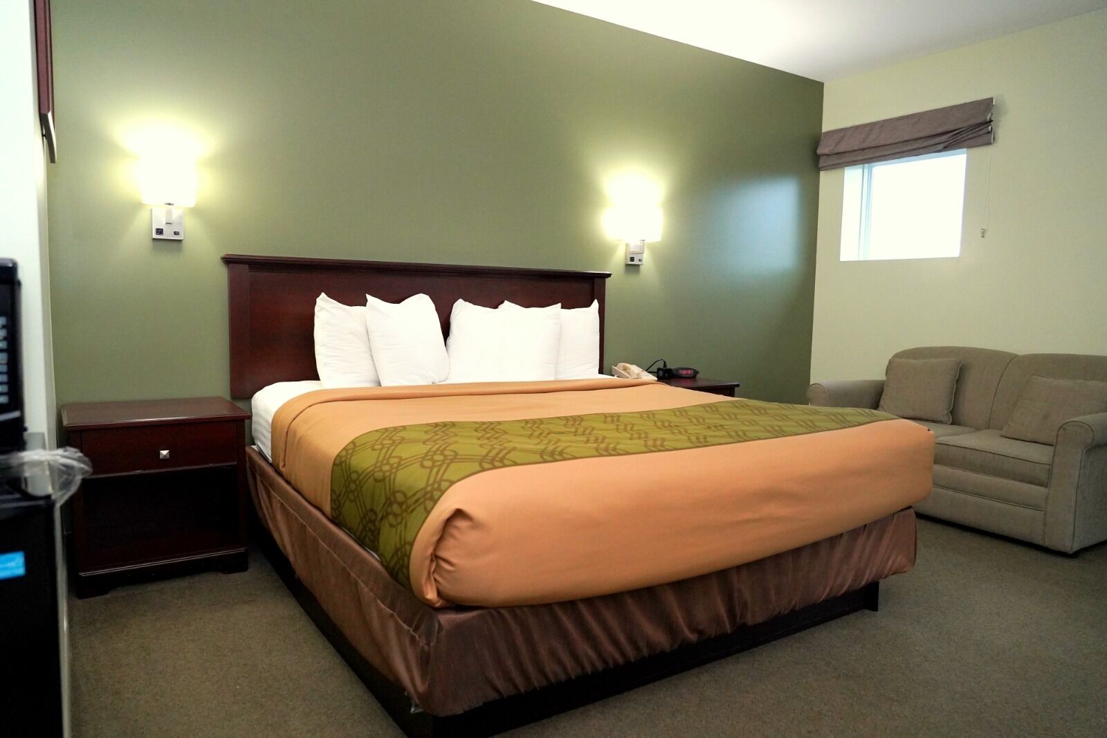 A hotel room with a bed, a nightstand, a lamp, a sofa, and a window with curtains.