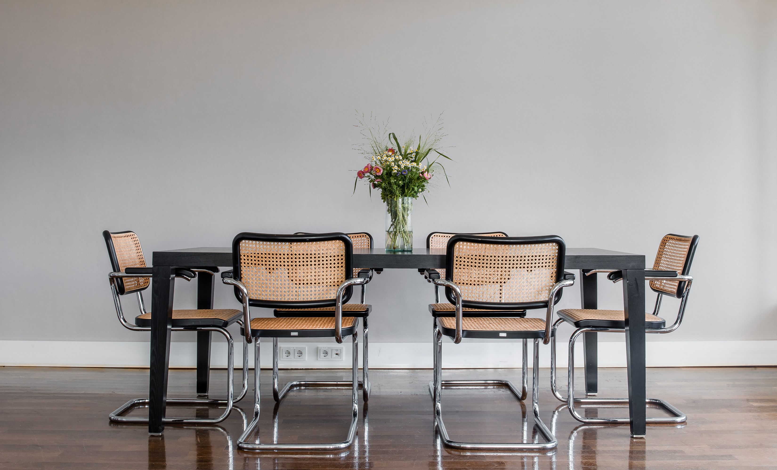 A dining table with four chairs, a vase with flowers, and a plain wall.