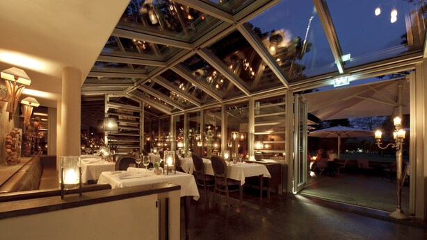 Un restaurante con techo de vidrio, mesas dispuestas con manteles blancos y vistas al exterior.