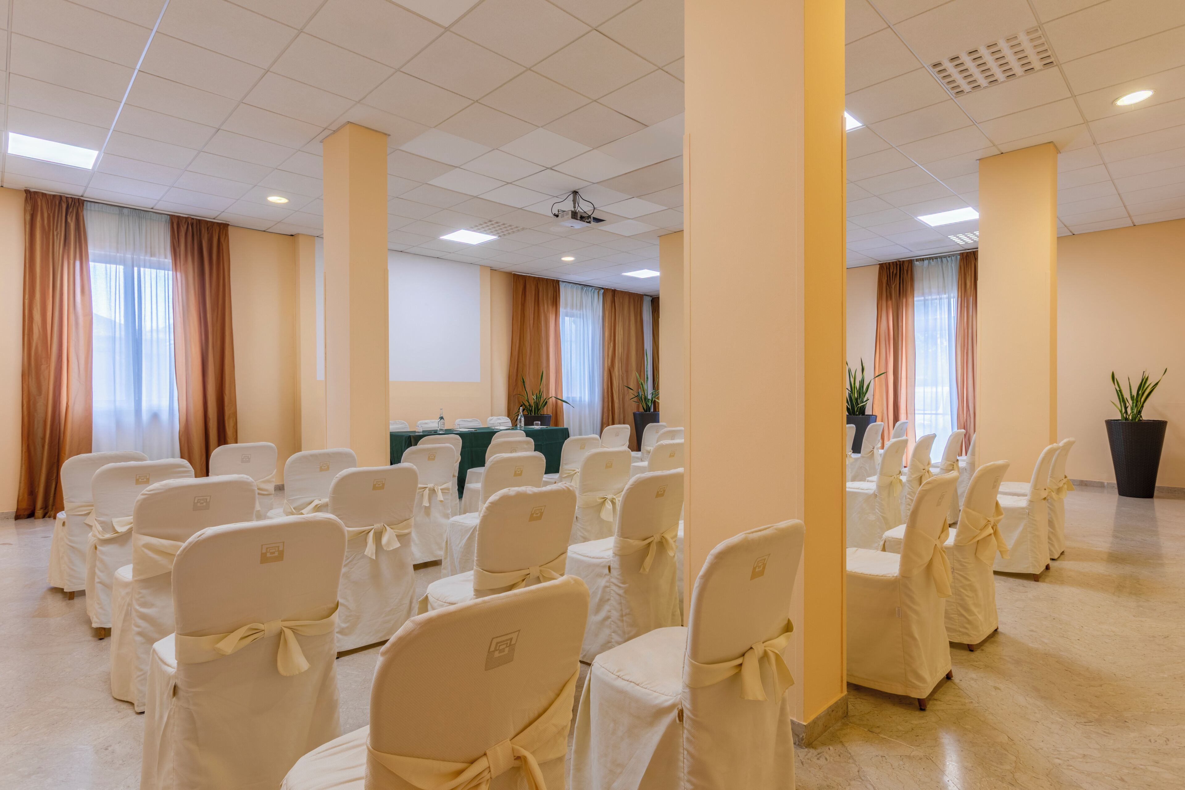 A conference room with rows of chairs, a table with a projector, and potted plants.